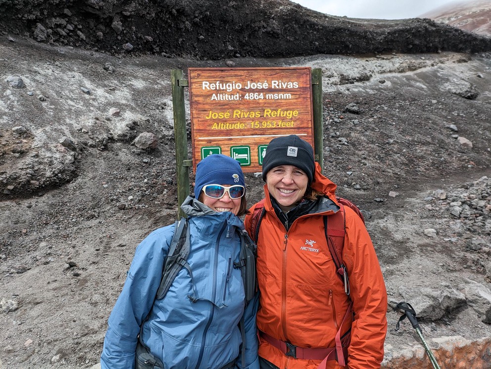 two women stand in front of a sign indicating they have made it to a refuge for climbers high on a volcano